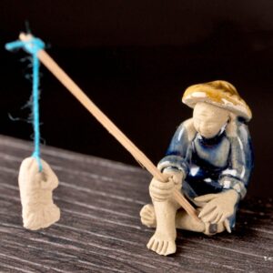 Tiny Ceramic Figurine of a Man Sitting Barefoot, Fishing with a Wooden Rod and a Hanging Shoe As Bait on a Dark Background